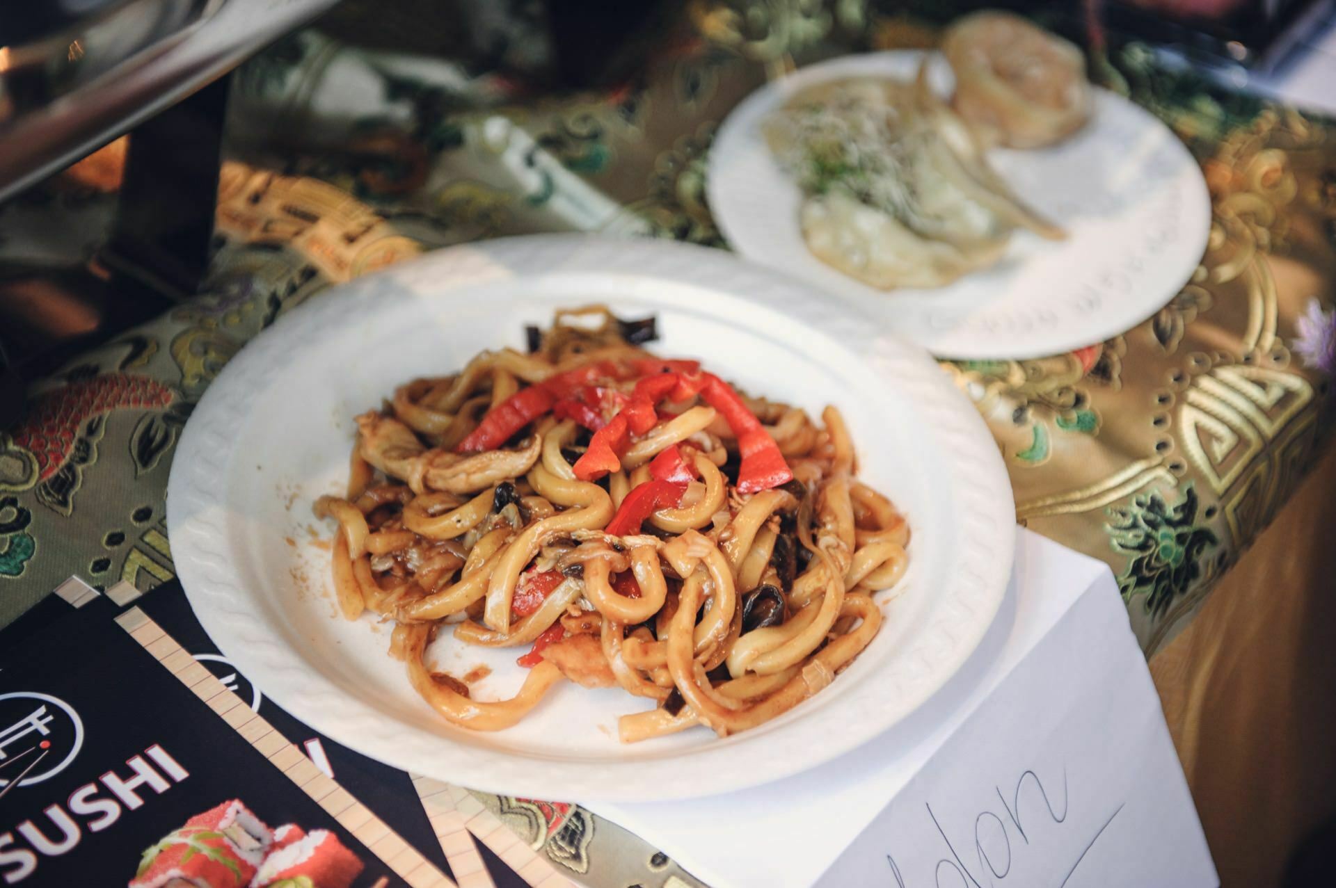 Close-up of a plate of udon noodles with red peppers and mushrooms, placed on a patterned tablecloth at a food fair. On the side a white plate with dumplings. A sushi menu is partially visible in the corner.  