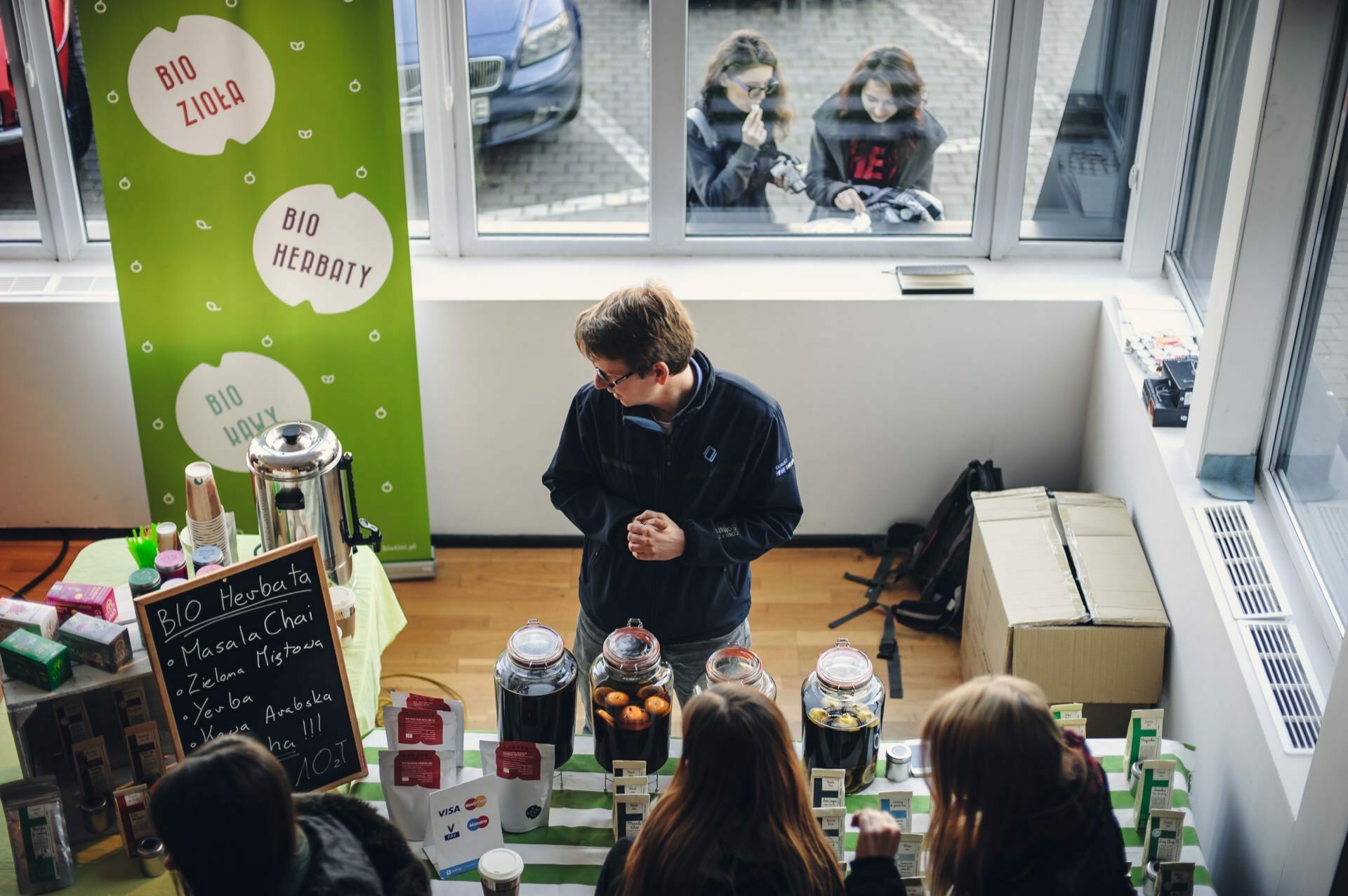 A vendor at an indoor booth stands behind a table on which jars of tea leaves and a dispenser of hot drinks are displayed. Served are three customers with their backs turned to the camera. Outside, two people are peering through a window, watching the bustling scene from the food fair.  