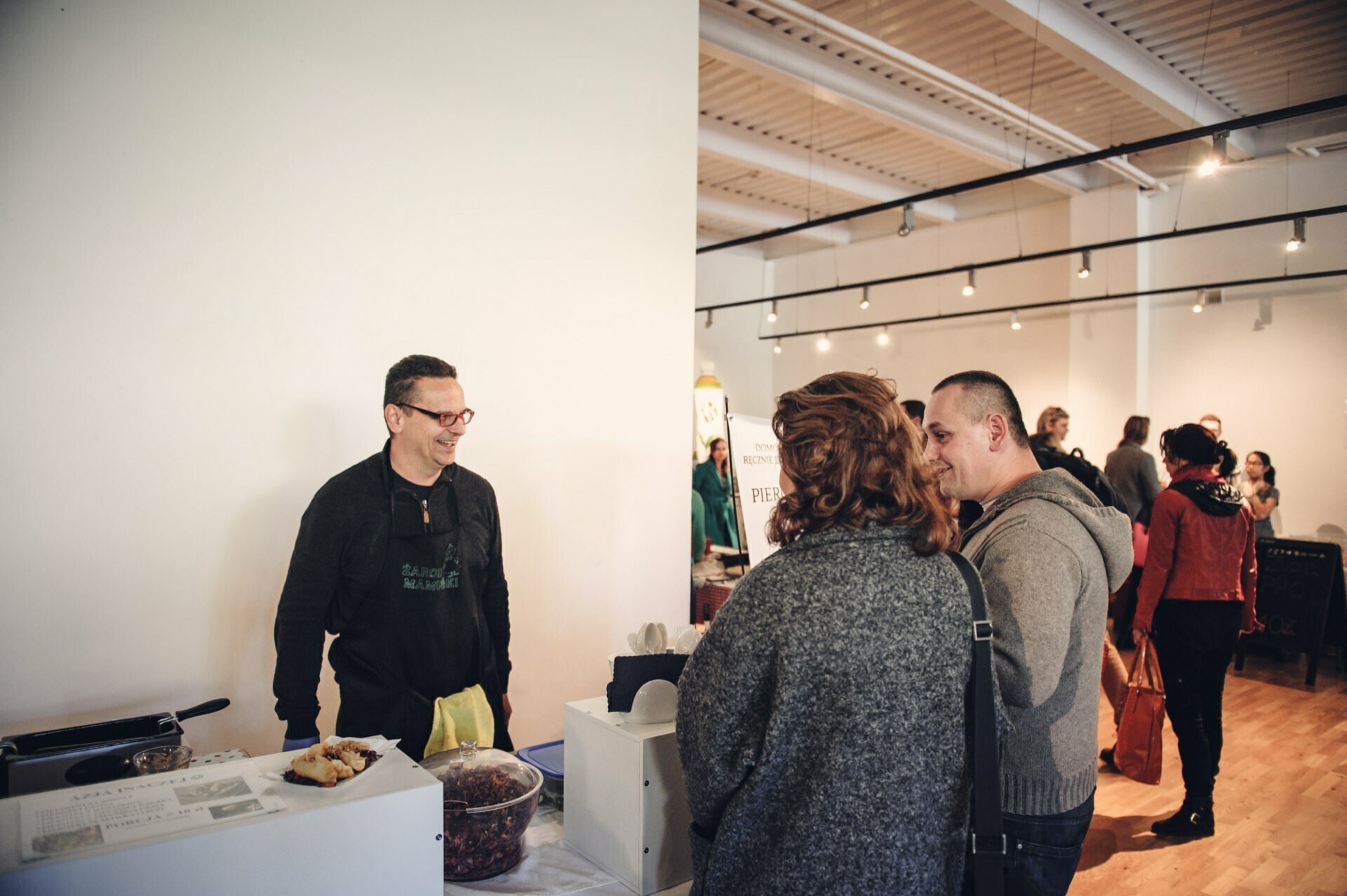 A man wearing glasses and an apron stands behind a table of food products, smiling and chatting with two people at a food fair. The event takes place in a large, bright room, with several people mingling in the background. The atmosphere seems casual and friendly.  