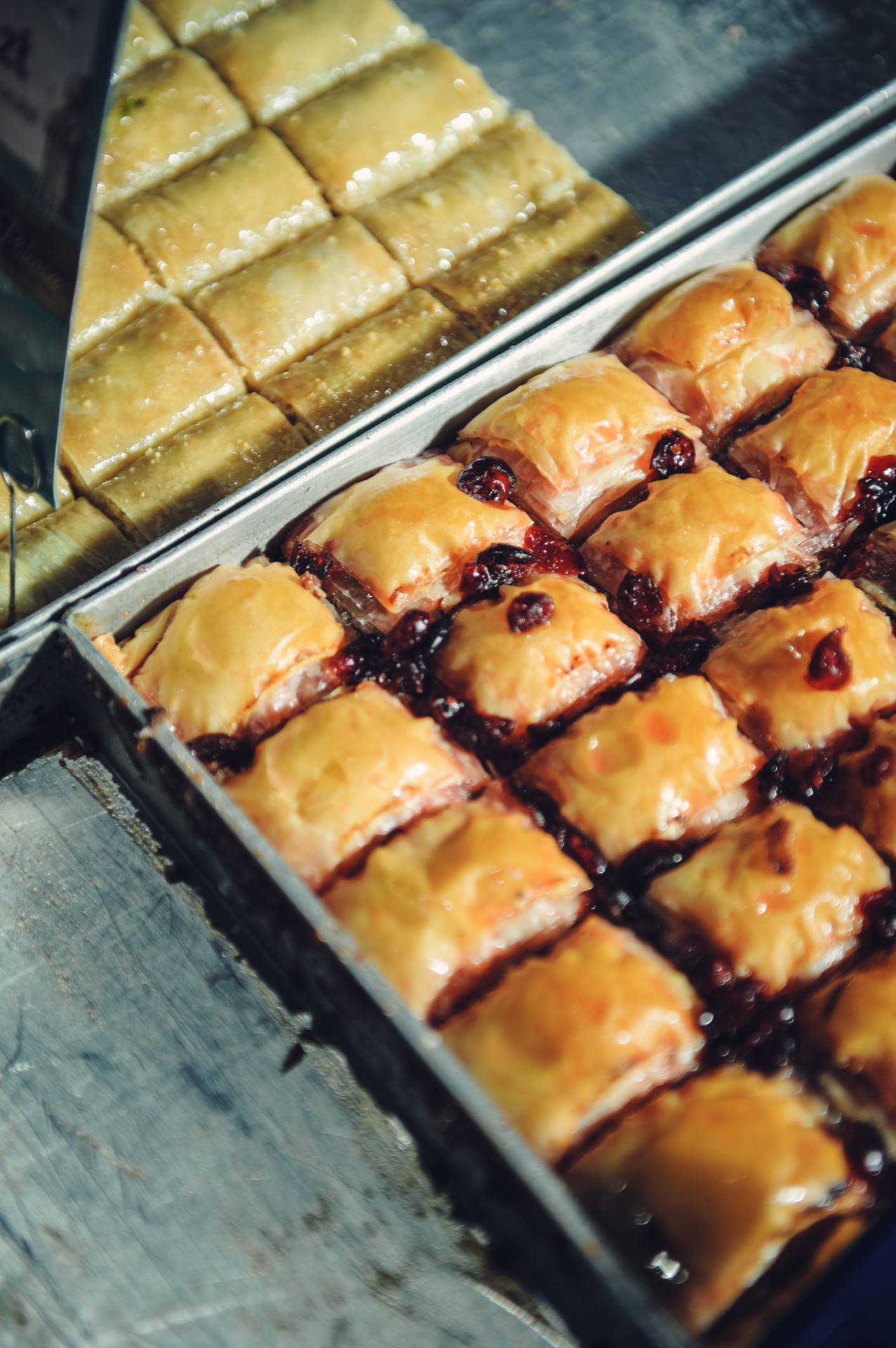 Two trays of baklava stand side by side. The tray in the foreground features pieces of baklava with a glossy golden brown finish and cherry sprinkles, while the tray in the background depicts plain baklava with a crisp, glossy exterior. The freshly baked treats make a great presentation at food fairs.  