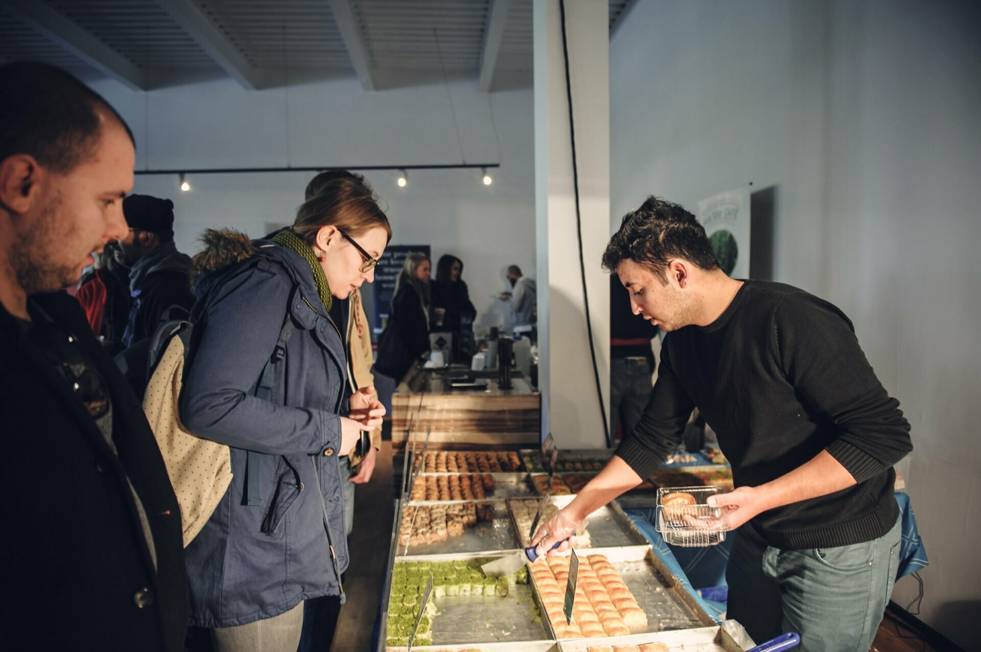 Customers are seen at a food stand, where one man is serving baklava and other baked goods. Two customers, one wearing a blue jacket and a backpack, carefully observe the offerings. In the background, other shoppers can be seen in the well-lit market space, highlighting the vibrant atmosphere of the food fair.  