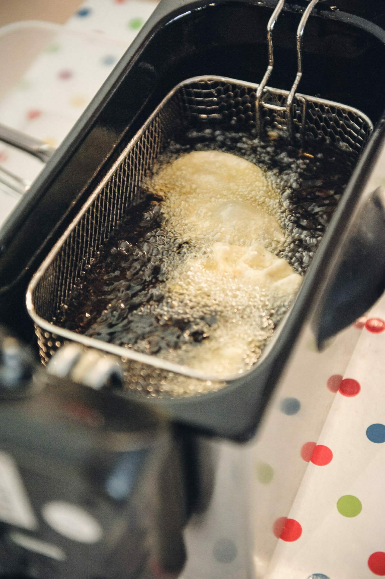 A hot oil fryer was shown at a cooking fair. The basket holds two pieces of batter that bubble as they cook. The fryer is mostly black, and a colorful dot pattern on the surface it sits on adds a cheerful touch.  