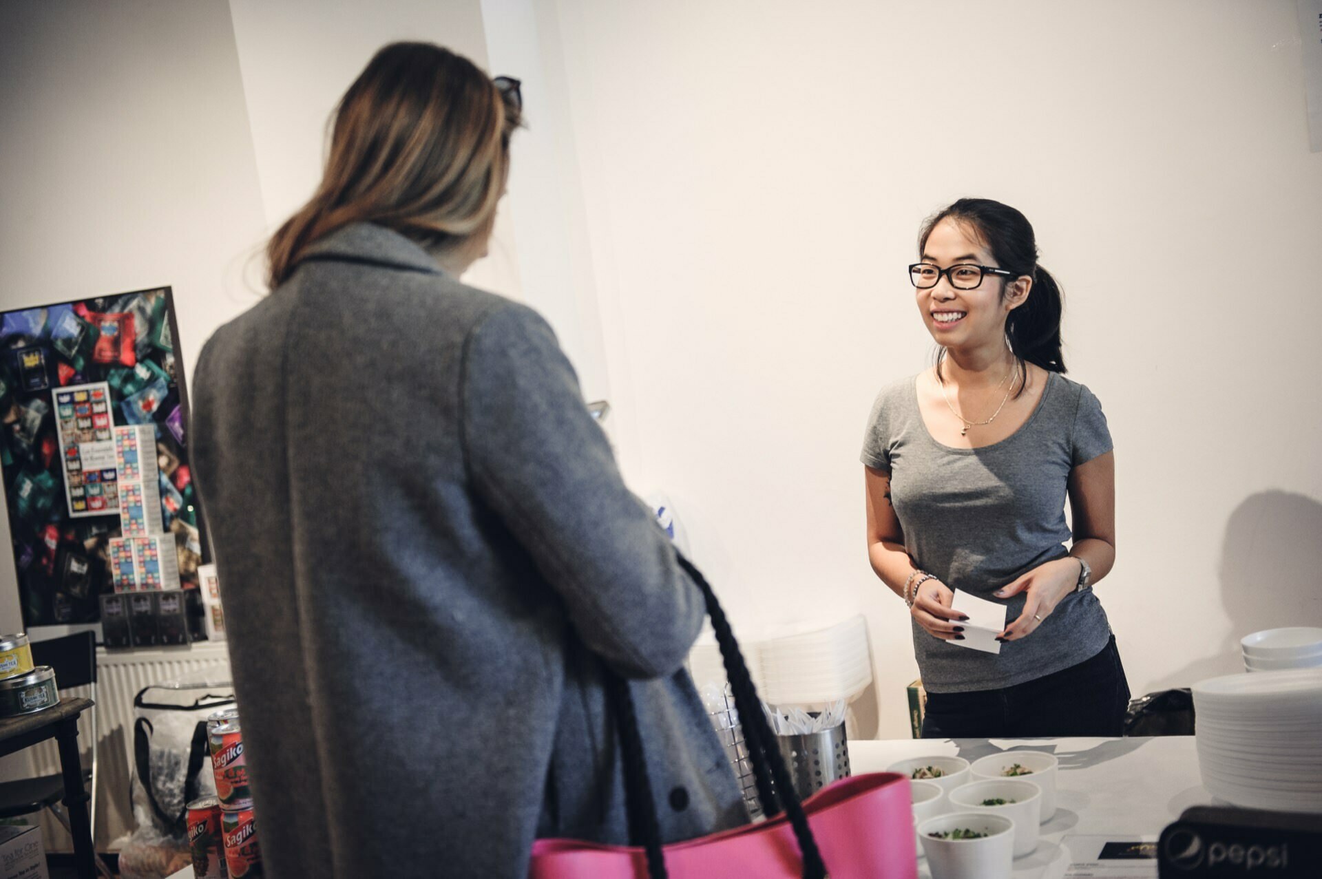 A young woman in glasses and a gray t-shirt stands behind a counter with bowls of food and smiles at another person in a gray coat holding a pink and black bag. The place resembles a casual restaurant or cafe, perhaps inspired by the lively atmosphere of a food fair. 