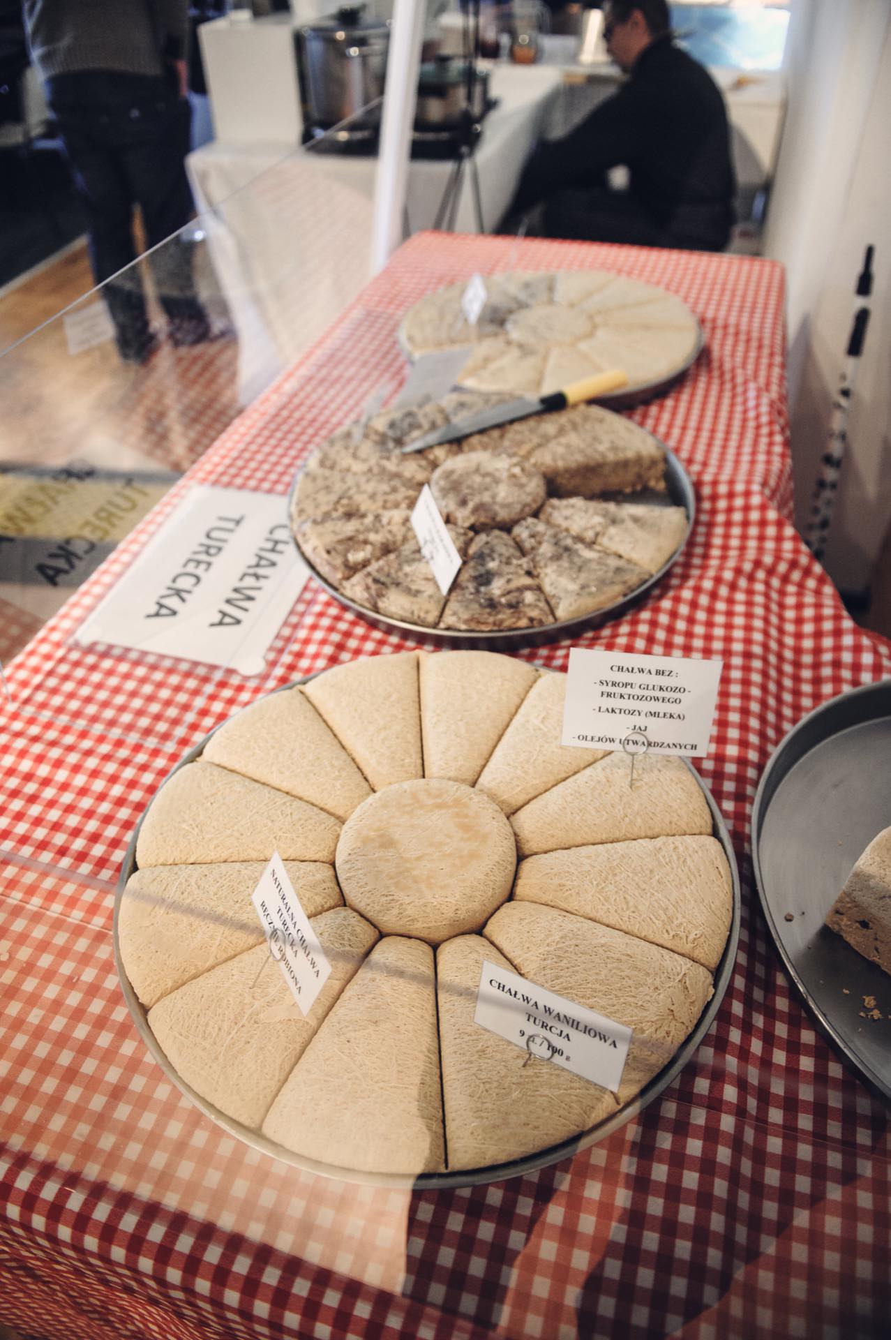 A display of various cheeses on a red and white checkered tablecloth, reminiscent of a culinary fair. Each cheese is marked with a plate indicating its name and origin. In the background you can see a somewhat blurry person working or examining something behind a transparent plastic barrier.  