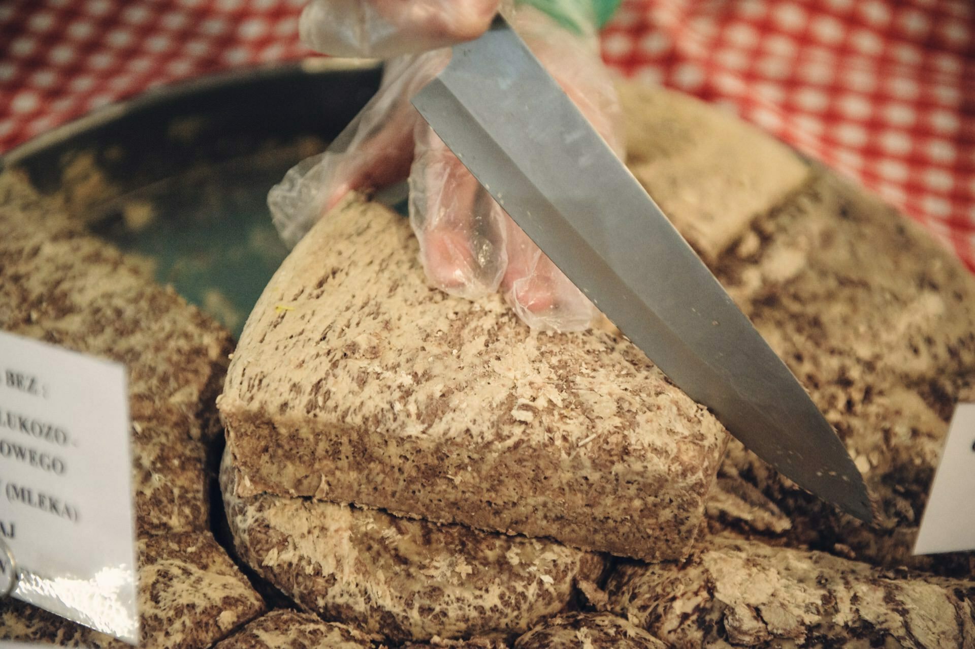 A plastic-gloved hand deftly slices a large, rectangular block of beige spotted cheese at a food fair with a large knife. The block rests on a pile of similar pieces of cheese, arranged on a red and white checkered cloth. The labels are visible but illegible.  