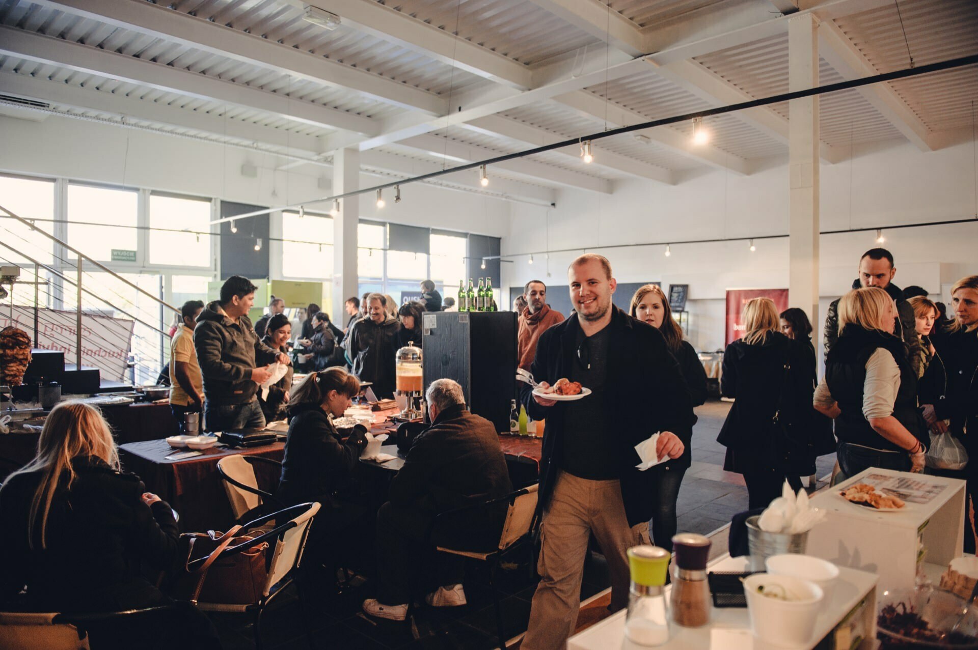 A lively indoor market, reminiscent of a bustling food market, with people gathered around tables and various stalls. Some sit, eat and talk, others stand or walk around. A man in the middle holds a plate of food and smiles. The atmosphere is lively and sociable.   