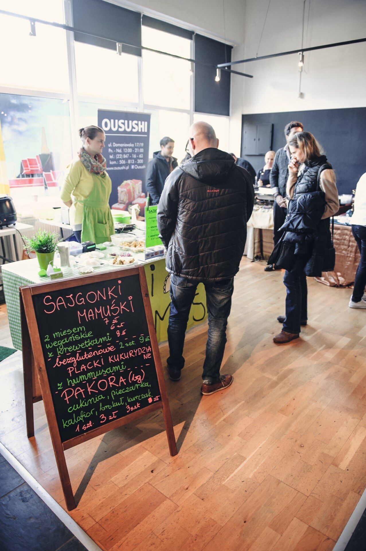 Several people stand at a food stand at the market, chatting and waiting to be served at the bustling food fair. In the foreground is a blackboard menu listing various dishes in Polish. In the background you can see more stalls and the "OUSUSHI" sign.  