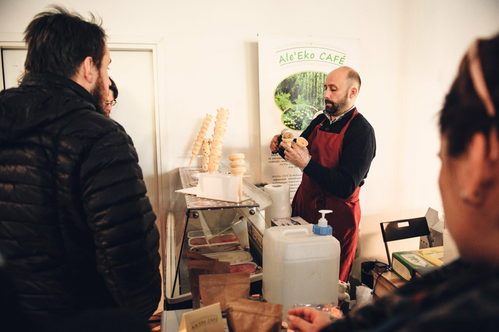 A man dressed in a red apron serves ice cream to customers at a booth labeled "Ale'Eko CAFÉ." The indoor booth, located at the culinary fair, consists of a table with various items such as a large white container, bags and a hand sanitizer pump. Three people gather around the booth.  