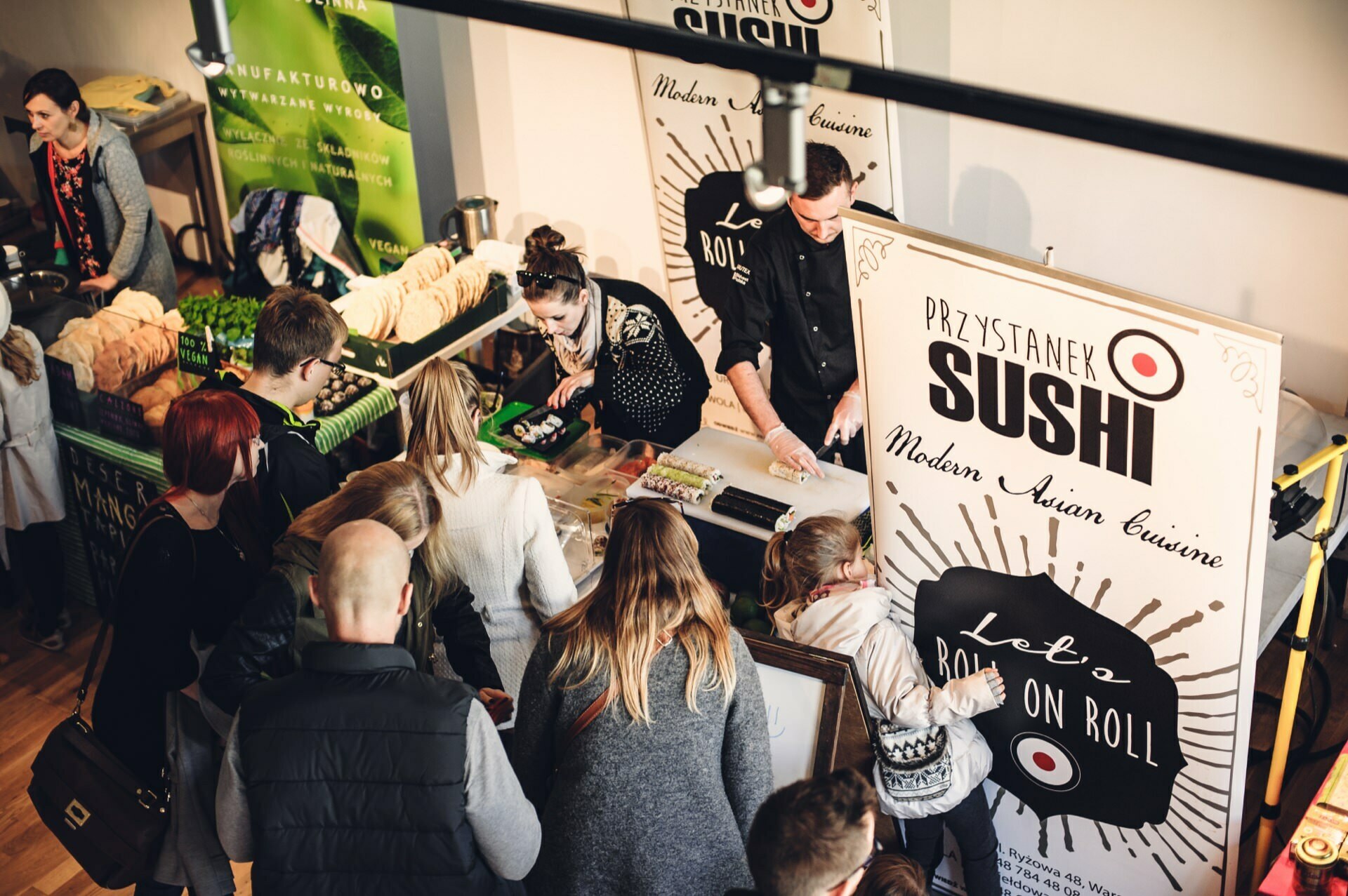 A crowd of people gathers around the "Sushi Stop" sushi stand at the culinary fair. The booth has signs advertising "Modern Asian Cuisine" and "Let's Roll on Roll." Staff behind the counter prepare and serve food, while other booths and attendees can be seen nearby.  