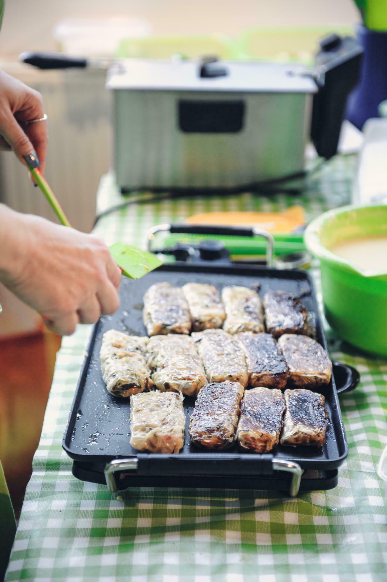 A person cooks many pieces of food, probably fish, on a black electric grill at a cooking fair. The grill is set on a table covered with a green and white checkered tablecloth. In the background is a green bowl and other kitchen objects.  