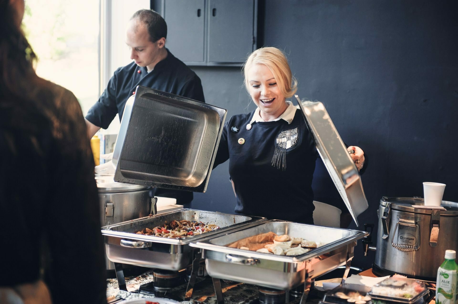 A woman smiles as she lifts the lids of two trays of various buffet-style dishes at a food fair. Behind her, a man, presumably a chef, is focused on preparing food. The place looks like a professional kitchen with cooking equipment and ingredients visible.  