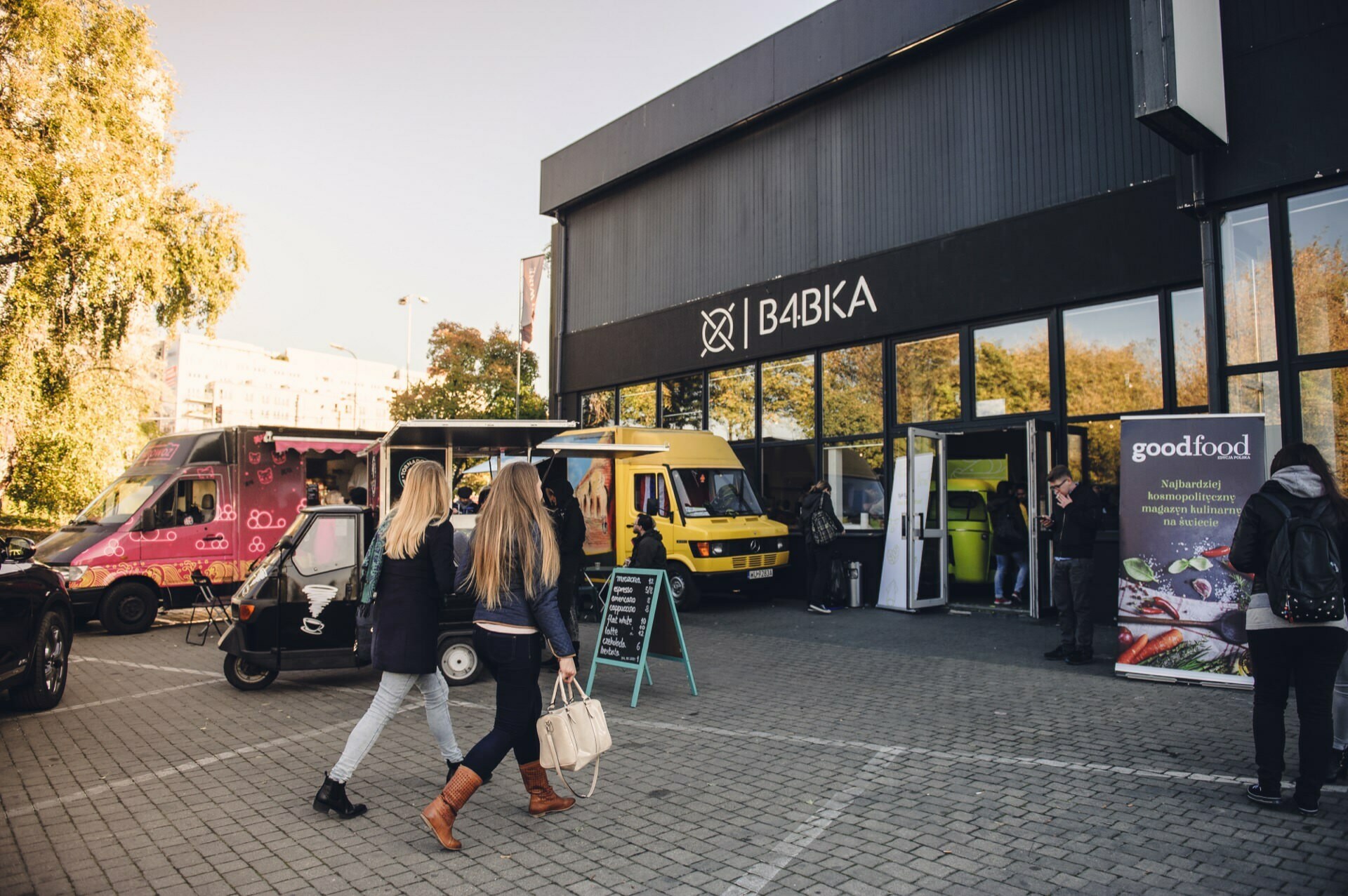 People strolling in front of the food market building with the sign "Babka". Various food trucks, such as pink and yellow, are parked outside. At the entrance there is a banner that says "good food". There is a lively and bustling atmosphere at these food markets.   