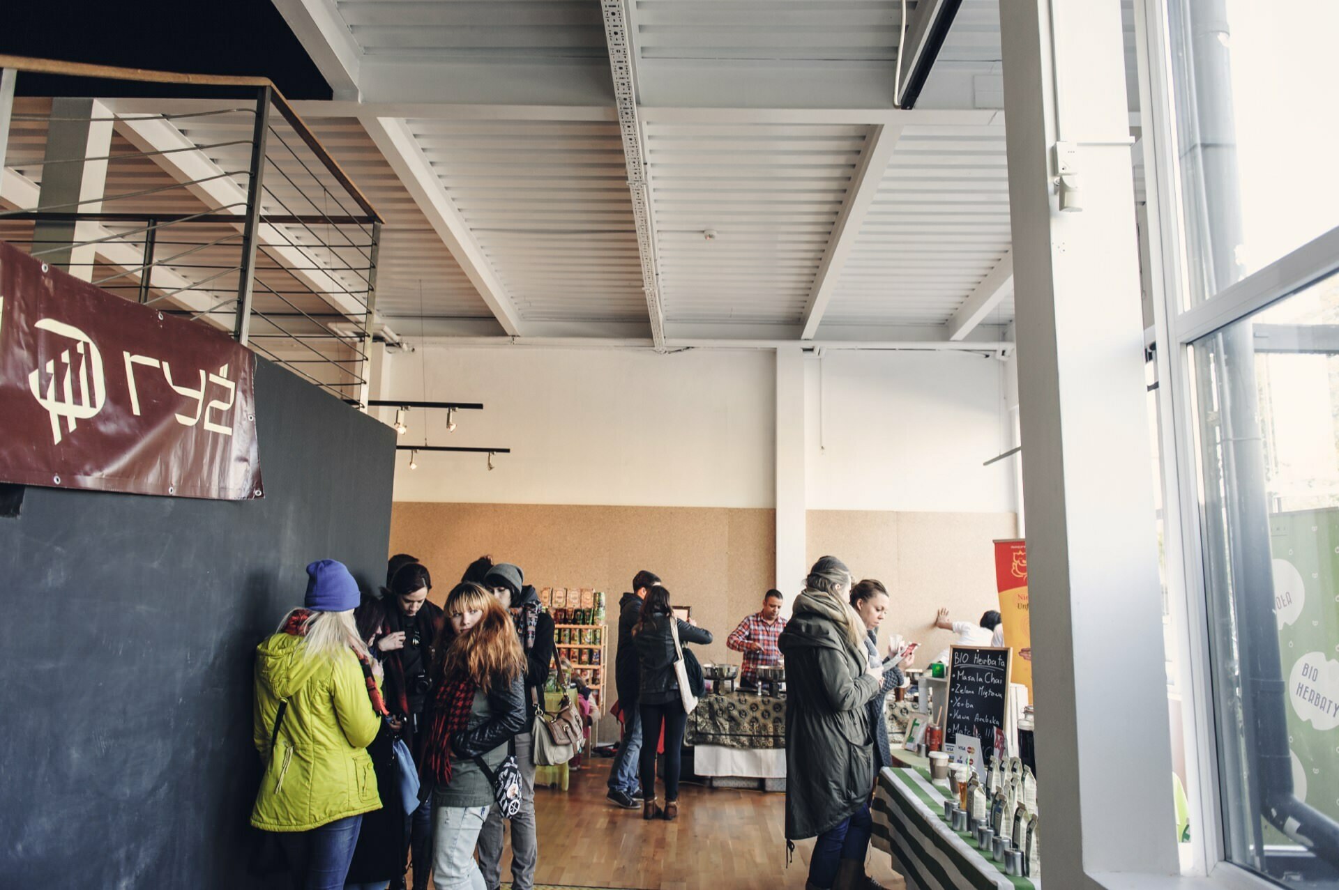 A group of people gather at a covered market, reminiscent of a food market. They browse and chat near various stalls filled with goods. The space has a modern, industrial look with large windows, white walls and exposed ceiling beams. A banner and shelves are visible in the background.   