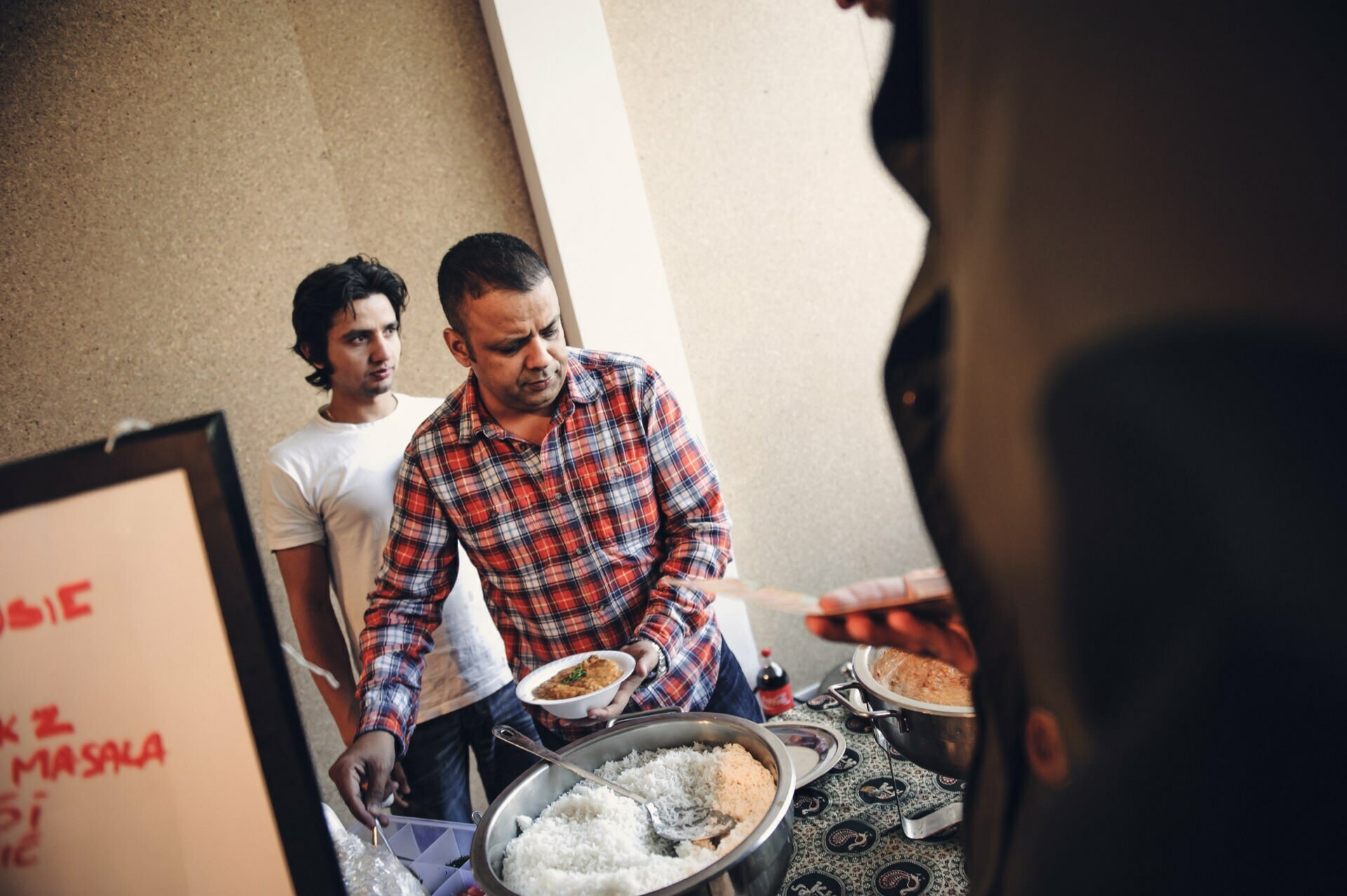 Two men stand behind a counter at a food market. The man on the left is wearing a white shirt and the man on the right, wearing a red plaid shirt, is serving food on a plate. In the foreground is a large bowl of rice and a second person holding the plate. A menu board is partially visible on the left.   