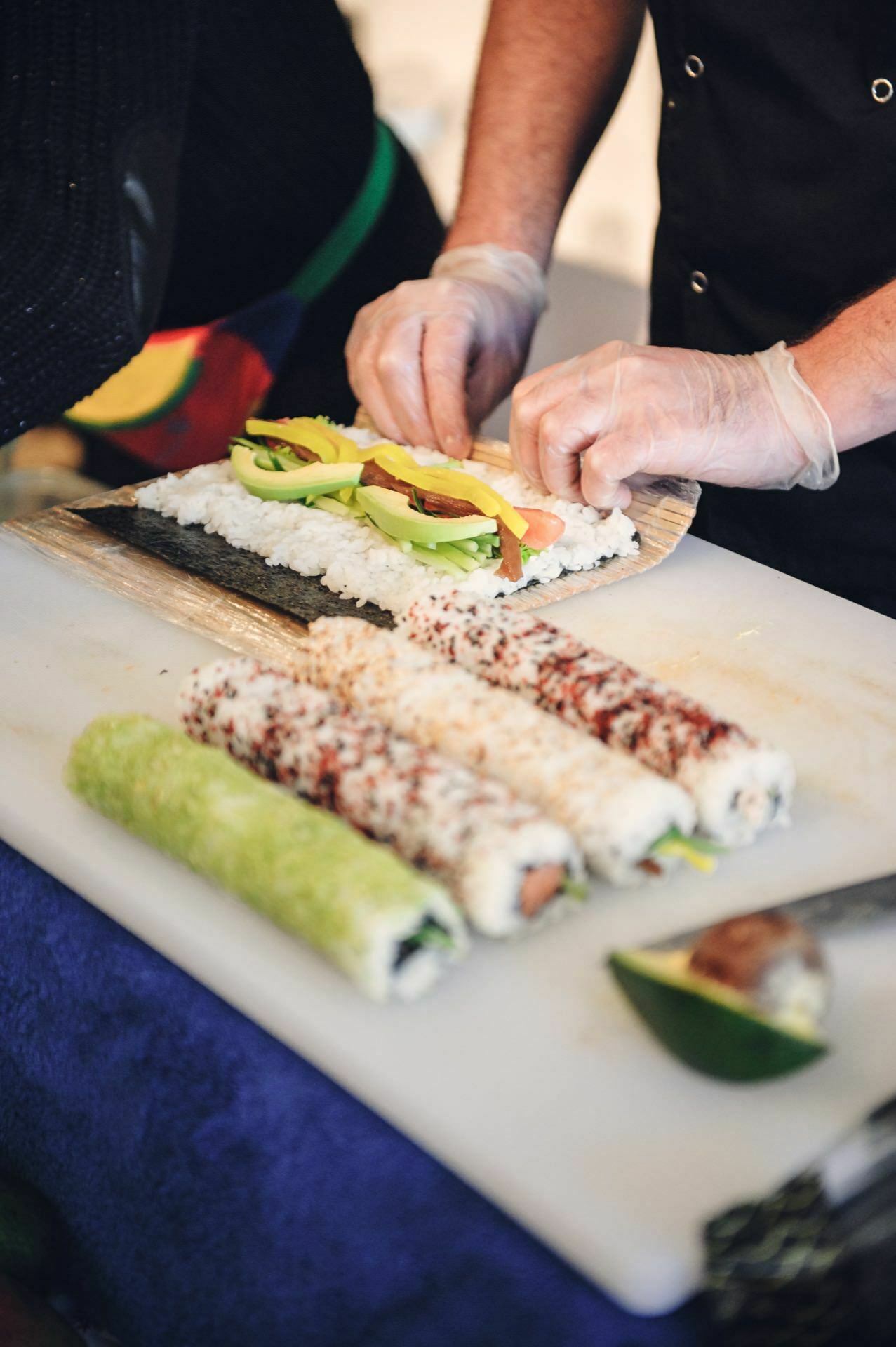 A person wearing gloves prepares sushi rolls on a white cutting board at a food fair. A sushi roll in progress contains slices of avocado on rice and seaweed. In the foreground, finished sushi rolls, one green and the other two covered with fish roe.  