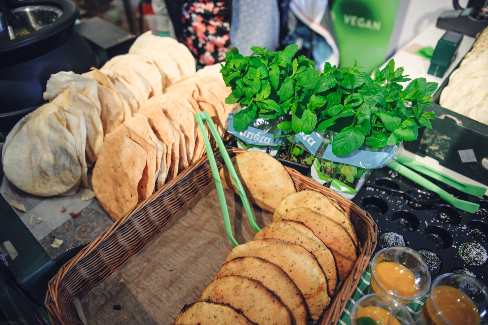 A display of fresh produce and food items. In a wicker basket are stacks of scones, several bunches of fresh herbs in green packages, small containers of orange-colored sauce and greens. A sign reading "Vegan" is visible in the background by the food market.  