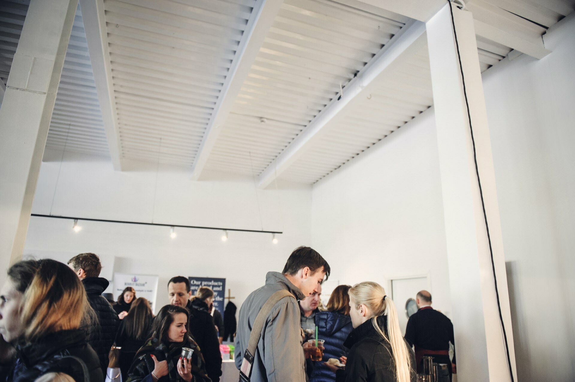 A group of people gather in a brightly lit, modern room with white walls and high ceilings. They are having conversations and drinks, presumably discussing the delicacies of the food fair. You can see posters and signs in the background.  