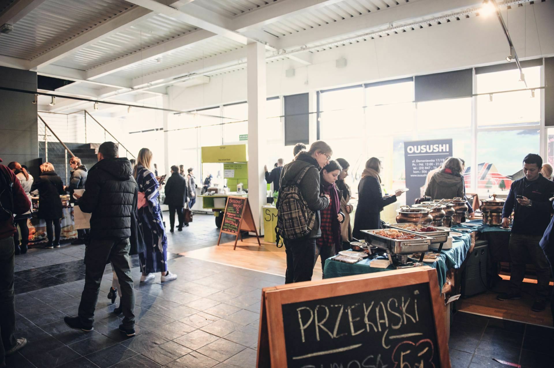 People huddle around a spacious, covered market, reminiscent of a bustling "food market," with various food stalls on display. In the foreground, a sign reads "FOREVER." Many people interact with the vendors, selecting food. Large windows allow natural light to fill the space.   