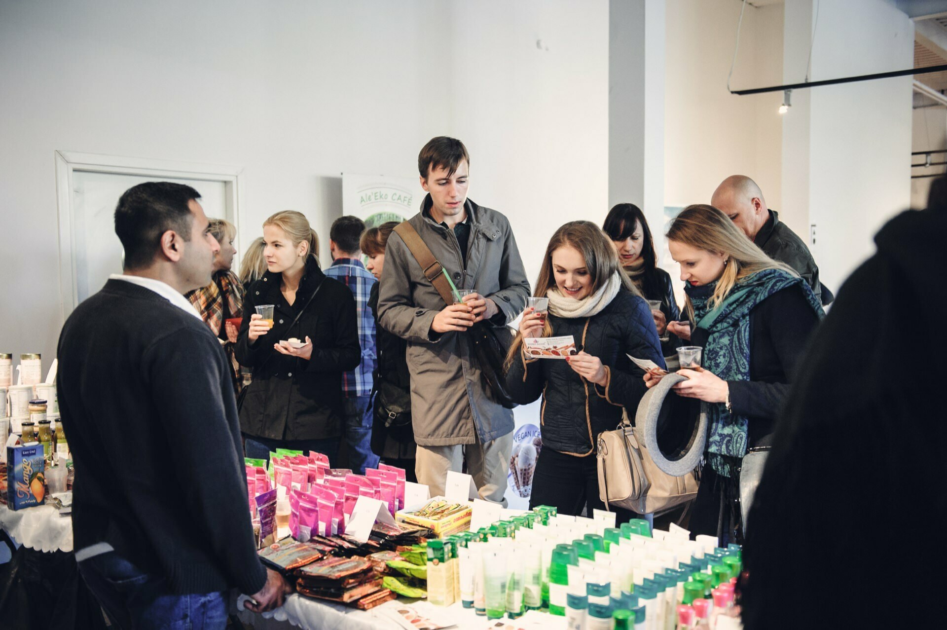 People gather around a table at a market or indoor market, looking at the various colorful products displayed on the table. Some hold the products, read labels or talk to each other. The scene at a food fair gives the impression of being casual and engaging.  