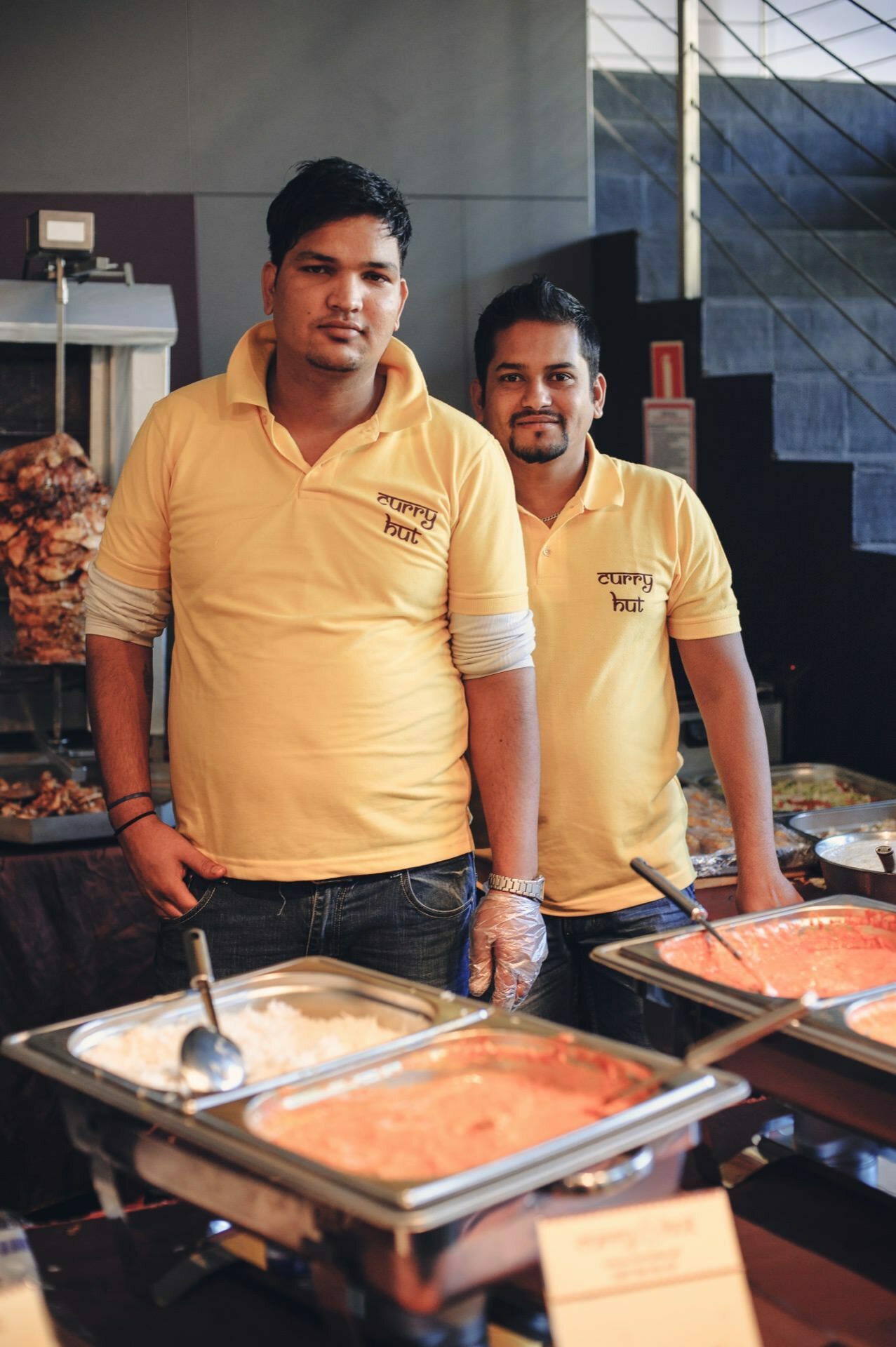 Two men in yellow shirts stand behind a counter with trays of various dishes, as if ready for a food fair. They are in an enclosed room with a metal staircase in the background. One man is holding a serving dish and both are smiling at the camera.  