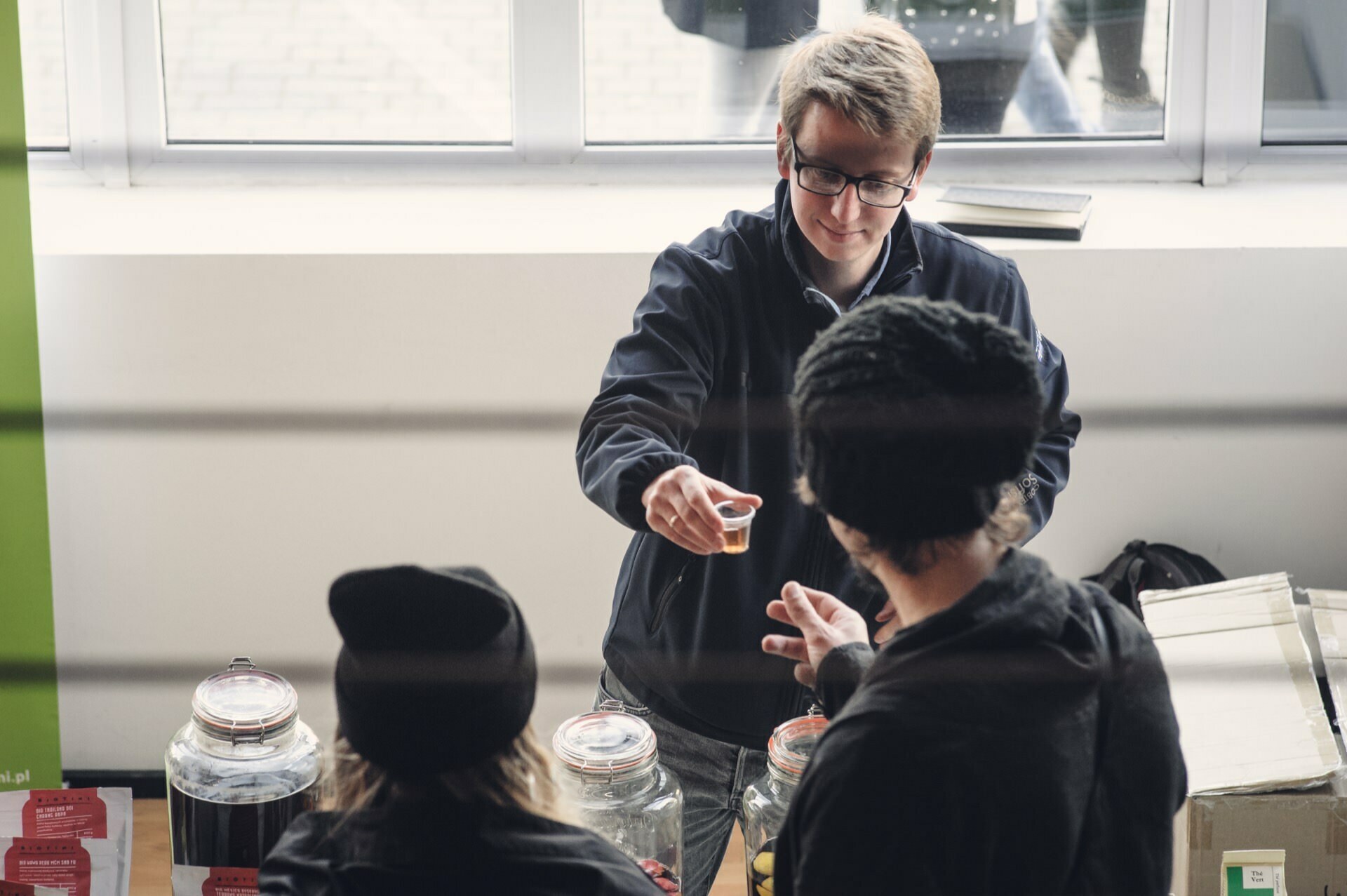 A person with glasses and short hair hands a small sample cup to two people in hats at a counter during a food fair. Large jars are stacked on the counter, and a window in the background lets the light shine through. 