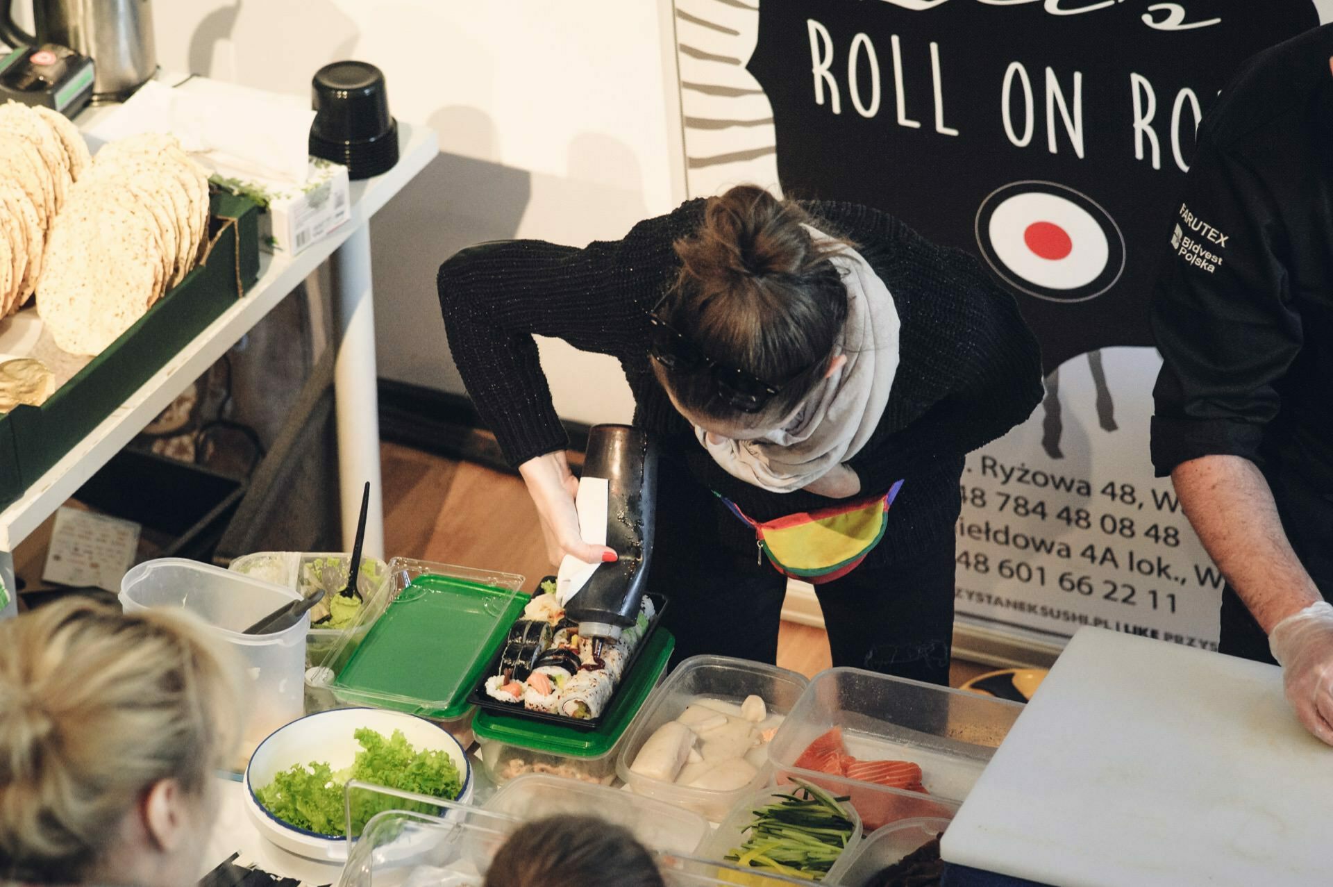 A person wearing a scarf and apron prepares sushi at a counter during a food fair. The countertop is filled with various fresh ingredients such as lettuce, fish and vegetables. In the background is a black poster that reads "Roll on Roll." Other people are nearby and watching.   