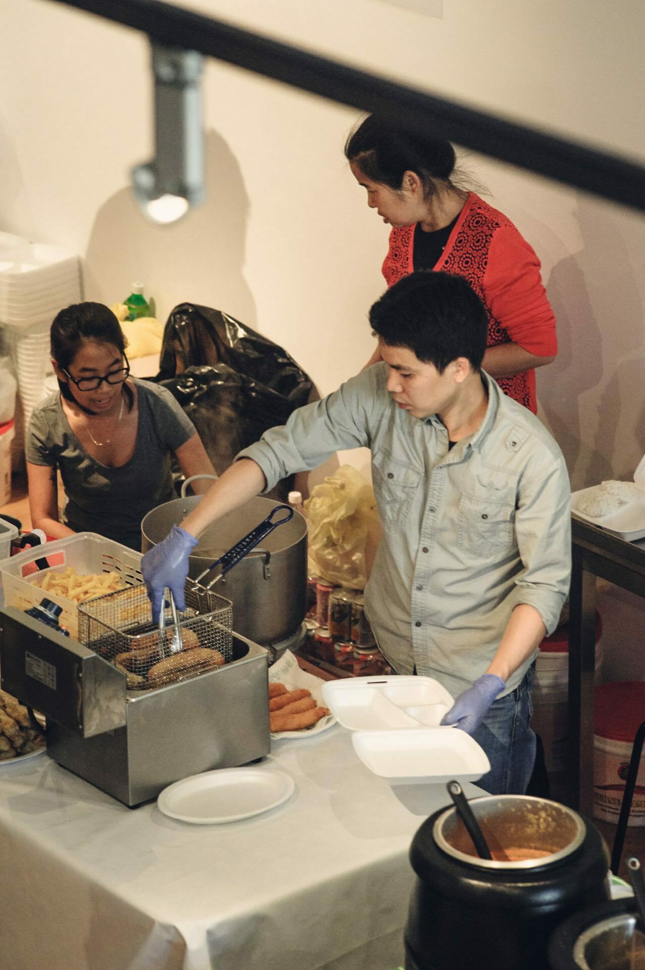 Three people are working in the kitchen. One man is using a deep fryer to prepare food, a woman next to him is holding a container, and another woman in the background is watching. Various kitchen utensils and ingredients are visible around them, giving the scene an atmosphere reminiscent of a food fair.  