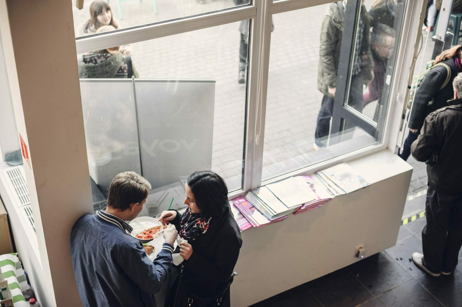 Two people are standing inside by a large window, sharing a plate and talking. Outside the window you can see several people passing by, perhaps going to a food fair. Inside, a stack of papers or magazines lies on a shelf by the window.  