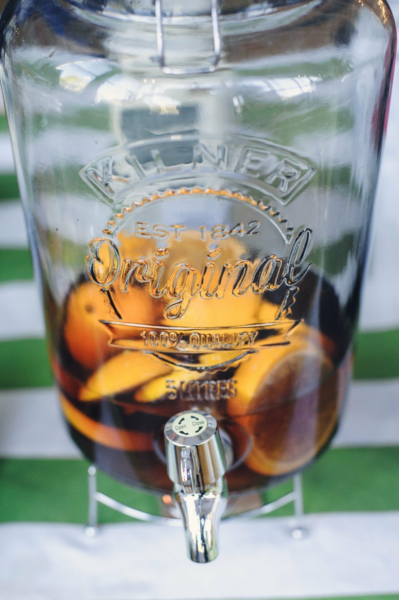 A clear glass beverage dispenser embossed with the words "Kilner Original, EST 1842," containing a beverage with lemon and orange slices visible. The dispenser, often seen at culinary fairs, features a shiny metal tap at the bottom and is set on a green and white striped surface. 