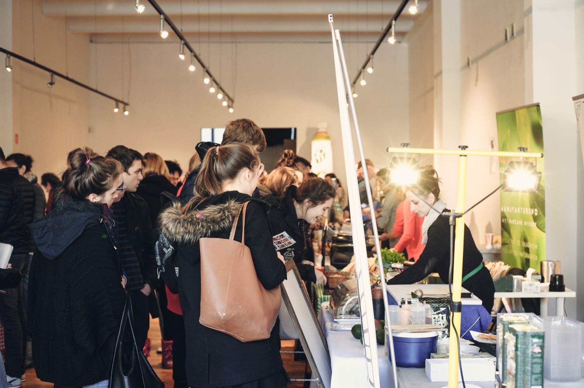 People gather in a bright indoor market. They are lined up at the stands of various vendors, and products are displayed on tables. Most people are wearing casual winter clothing, and one woman carries a brown bag. In the light of the overhead lamps, the atmosphere is that of a culinary fair.   