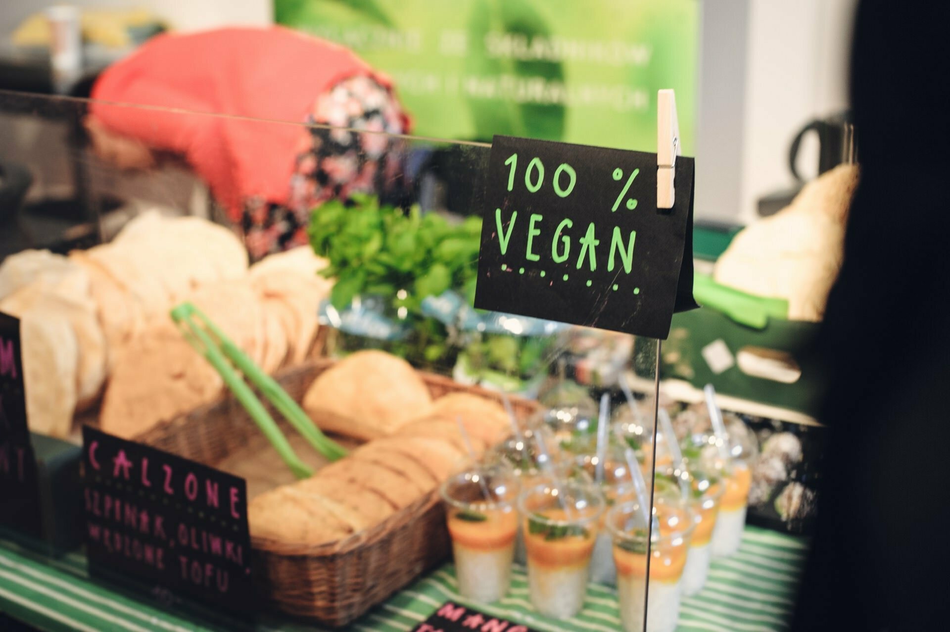 The table at the food fair showcased a variety of vegan dishes, including bread in a basket, green smoothies in plastic cups and several other items. A "100% Vegan" sign is prominently displayed. Green plants and other foods are visible in the background.  