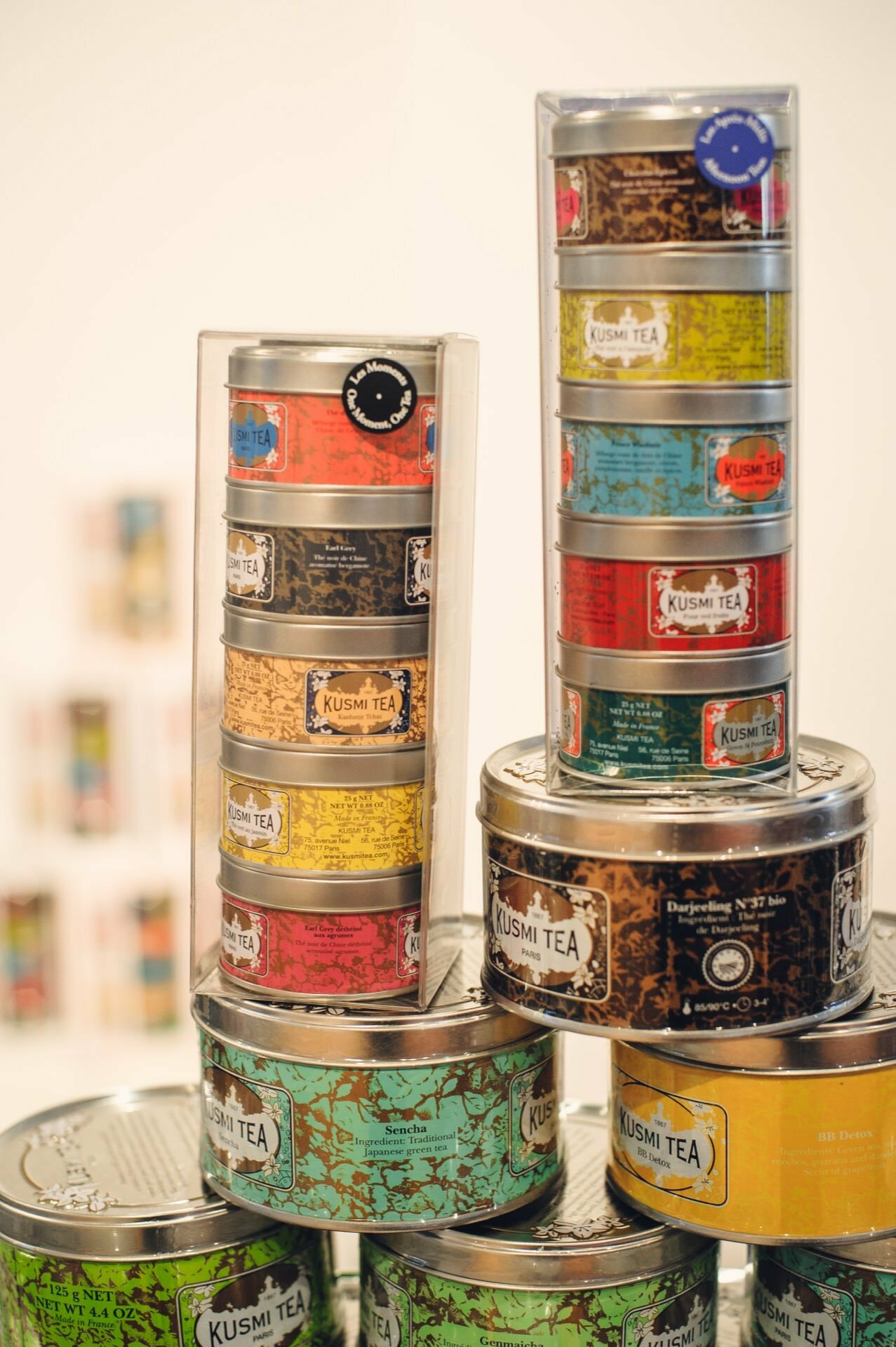 The photo shows a display of Kusmi Tea tins arranged in colorful arrangements at a food fair. There are two tall, clear plastic containers holding many small cans, and several larger cans are placed around them. The cans have different colors and designs, highlighting the brand's diversity.  