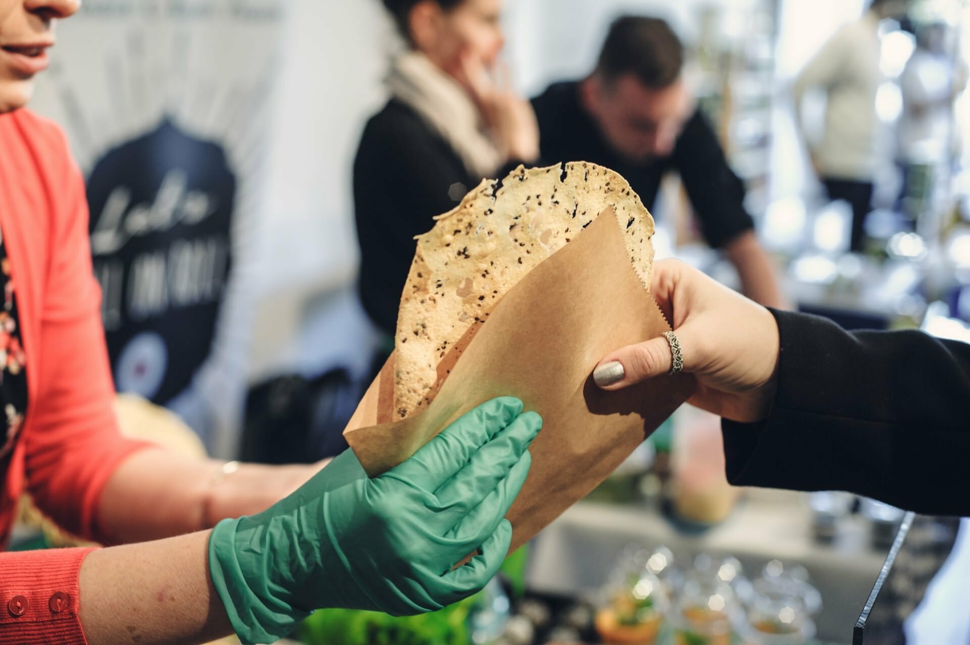 At a food fair, a person wearing green gloves hands a piece of crispy scones wrapped in brown paper to another person, who extends his hand to accept it. In the background you can see a blur of two people focusing on preparing food at a covered booth. 