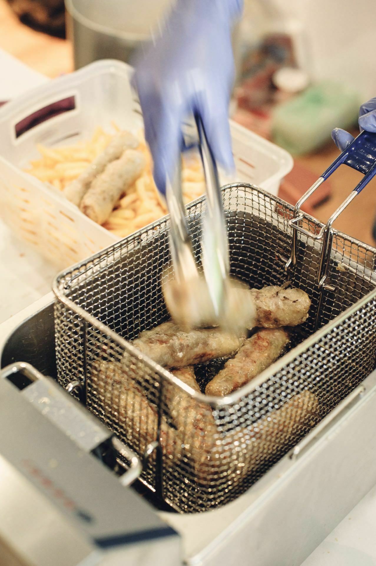 A person wearing blue gloves lifts a piece of fried food with tongs from a fryer basket filled with several pieces. A container of fries can be seen in the background. The environment, reminiscent of a food fair, resembles a kitchen or commercial food preparation area.  