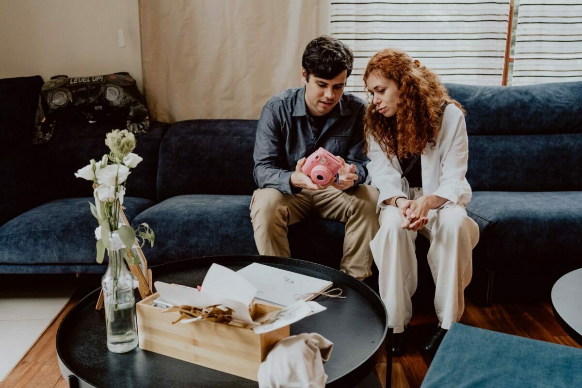 A man and a woman sit on a dark blue sofa and stare intently at a pink instant camera. In front of them is a black round coffee table with a wooden tray with papers and a tablecloth and a vase with white flowers. In the background is a window with striped blinds - another intimate shot by Marcin Krokowski, a leading photographer in Warsaw.  