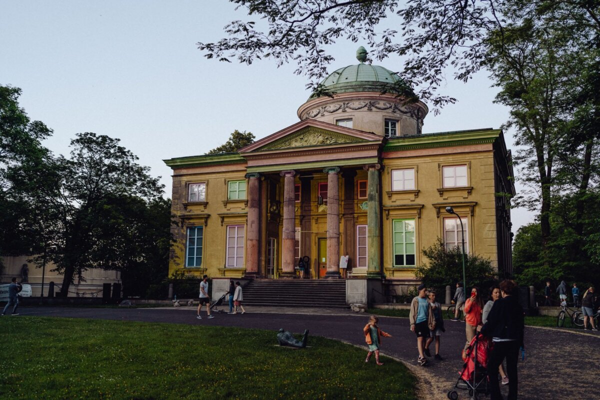 A neoclassical building with a dome and tall columns stands surrounded by greenery. People walk, run and push carts along the path in front of the building. The action takes place in the late afternoon, when the building is illuminated by soft sunlight, perfect for a photography event.  