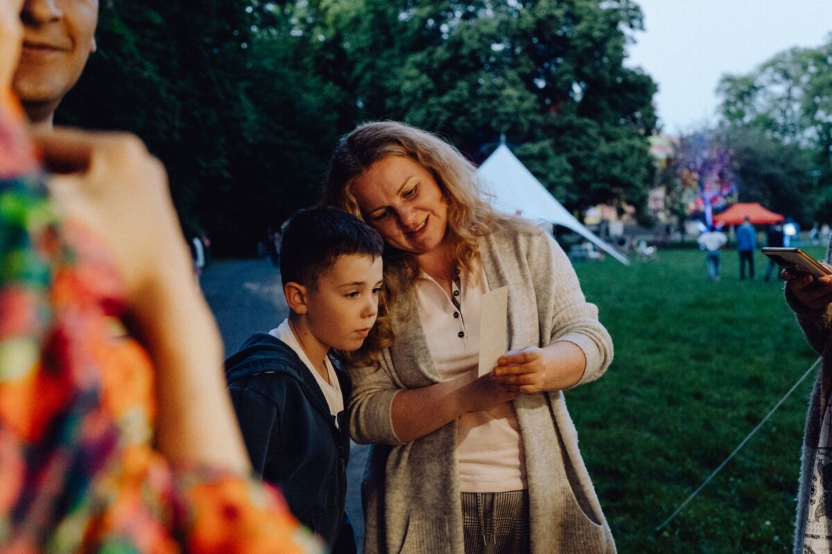 A woman with shoulder-length hair smiles warmly as she holds up a piece of paper for the young boy beside her to see. They are standing outdoors in a park with lush green trees and a white tent in the background, capturing a delightful photo moment of the event. Other people are partially visible around them.  