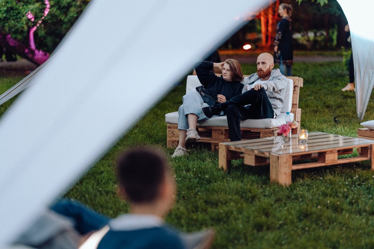 Two people are sitting and relaxing on a wooden outdoor couch with cushions, under a white canopy. In front of them is a low wooden table with a vase and a drink. In the background are trees and other faintly visible people, beautifully captured by event photographer Warsaw, creating a peaceful evening atmosphere.  