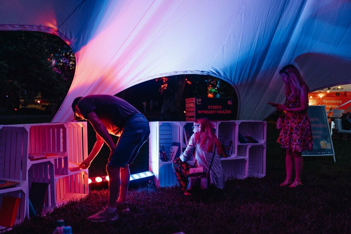 People browse through books in an open-air book exchange tent at night, illuminated by colored lights. The tent features white shelves filled with books. One person leans over to look at a book, another kneels on the ground, and another stands checking his phone - the perfect scene for a photographer's event on.  