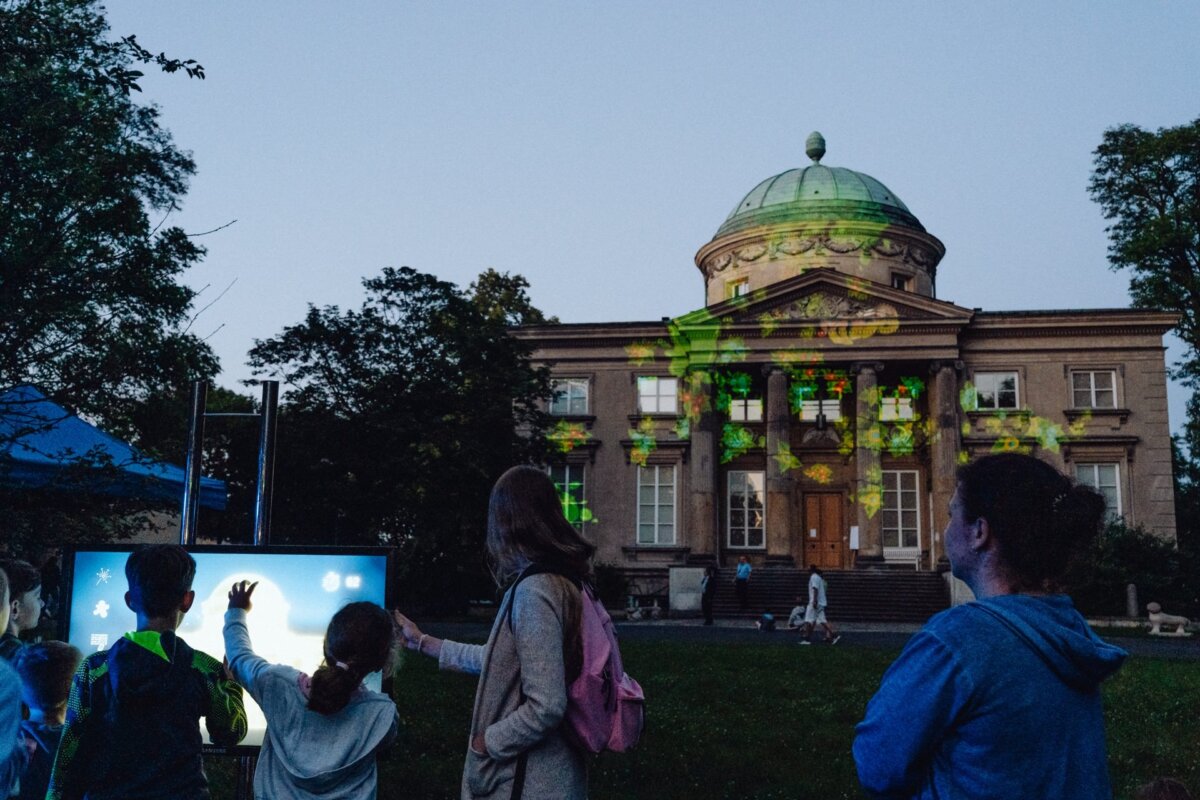 At dusk, people interact with an outdoor digital screen while a historic building is visible in the background with green and yellow patterns illuminated. Trees surround the scene, suggesting a community event captured by a talented photographer for the event, offering a vivid photo report of the event. 