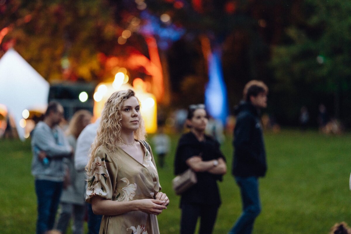 A woman with curly blond hair stands on a grassy area during what appears to be an outdoor event. Dressed in a floral dress, she looks off into the distance, capturing the essence of the event's photojournalism. Other people are casually gathered in the background, and colorful lights illuminate the scene.  