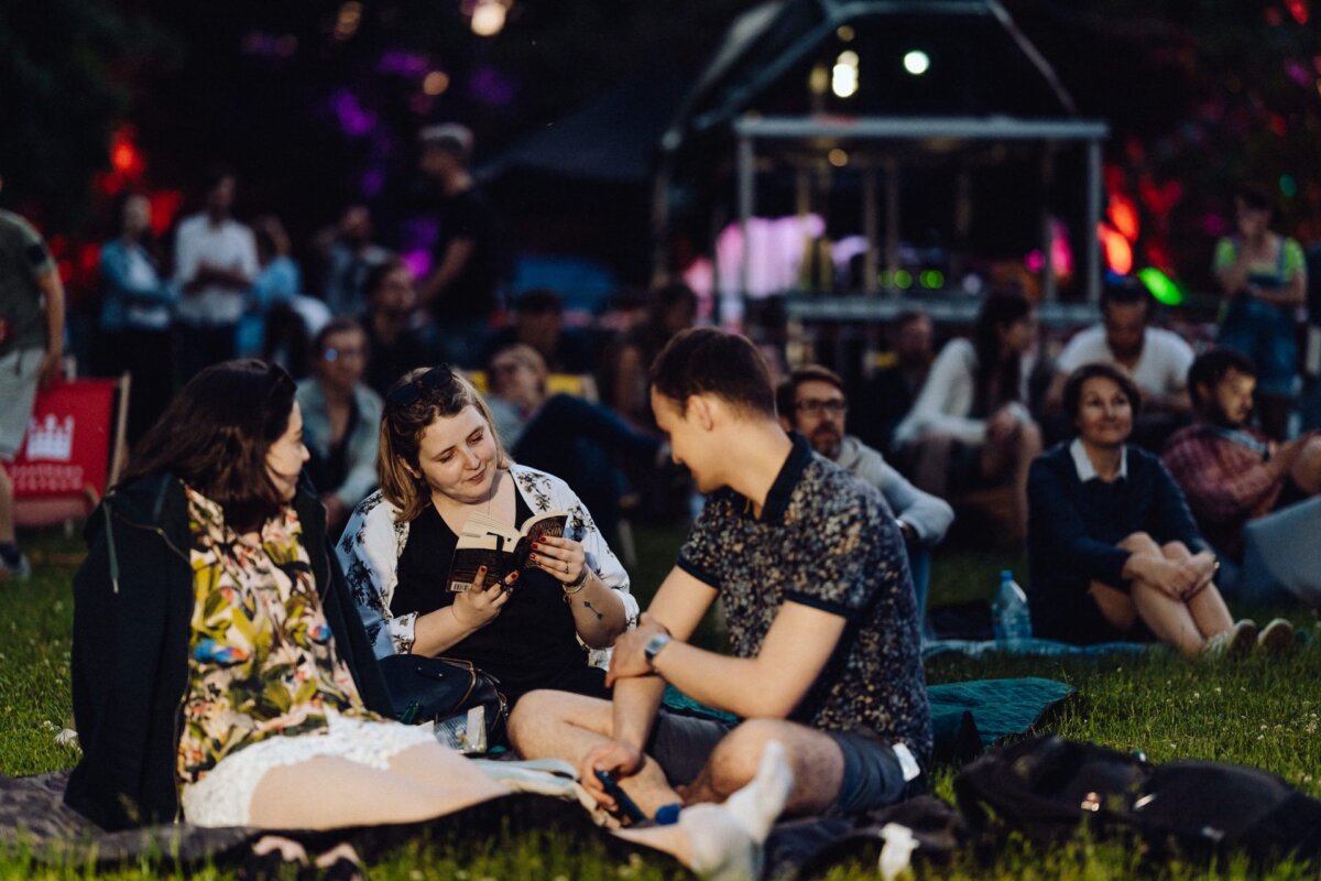 A group of three people are sitting on picnic blankets on the grass at an outdoor event. The person on the left is wearing a floral outfit, the person in the middle is reading a book, and the person on the right is engaged in conversation. Other participants and lights are visible in the background, captured in a photo essay of the event.  