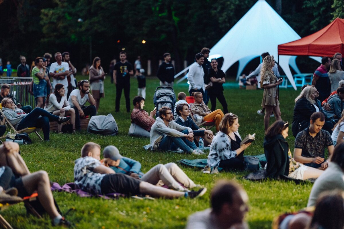 A diverse group of people sit and lie on the grass, face down, during an outdoor event held in the park in the evening. Some are lying on blankets or beanbags, others are standing or walking. In the background, a large white tent and a red canopy create a picturesque photo scene of the event.  