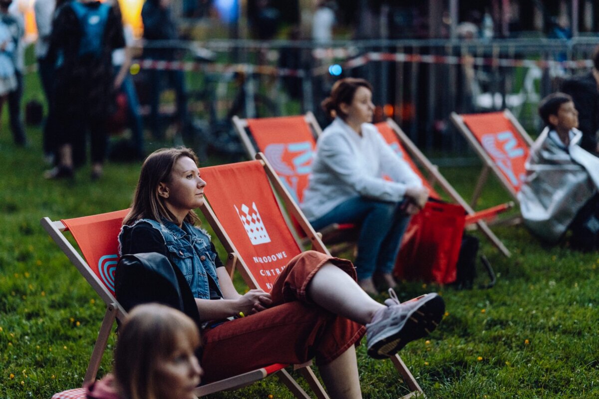 People are sitting on orange deck chairs in a grassy outdoor setting. They look relaxed and attentive, some wearing headphones. There's a festive atmosphere, and a few fuzzy figures near the railings add liveliness to the scene - a perfect photo-op of the event.  