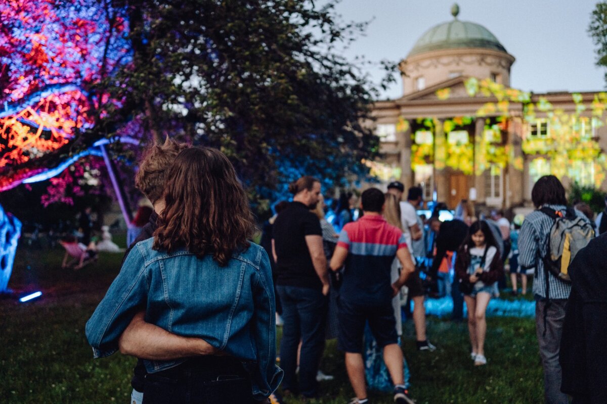 People gather outdoors near a building with a dome, illuminated by colorful lights cast on the trees and facade. A couple stands close to each other while others walk and interact in a relaxed atmosphere. The scene is festive and lively, perfect for a photo essay of the event.  