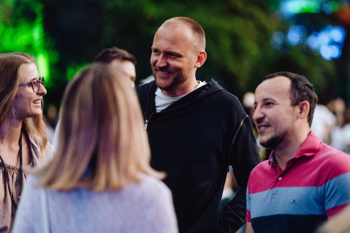 A group of four people are having a conversation outdoors at a social event. One man in a black hooded sweatshirt stands in the middle of the group and smiles. A blurred background, alluding to the greenery and colorful lights, adds to the charm of the event photo.  