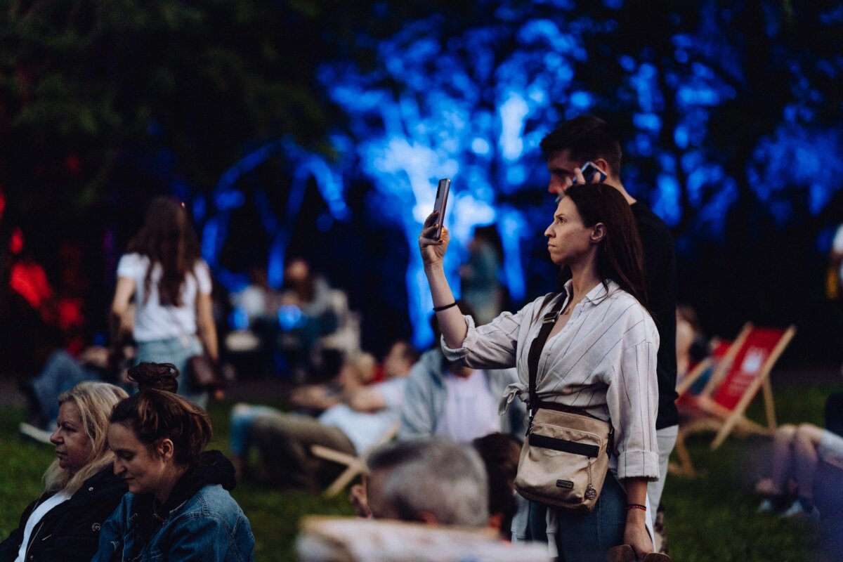 A woman stands up and picks up her phone, taking a photo report of the event in a park full of people, some sitting on the grass or on chairs. The scene is lit with colorful blue and red lights in the background, creating a festive atmosphere. 