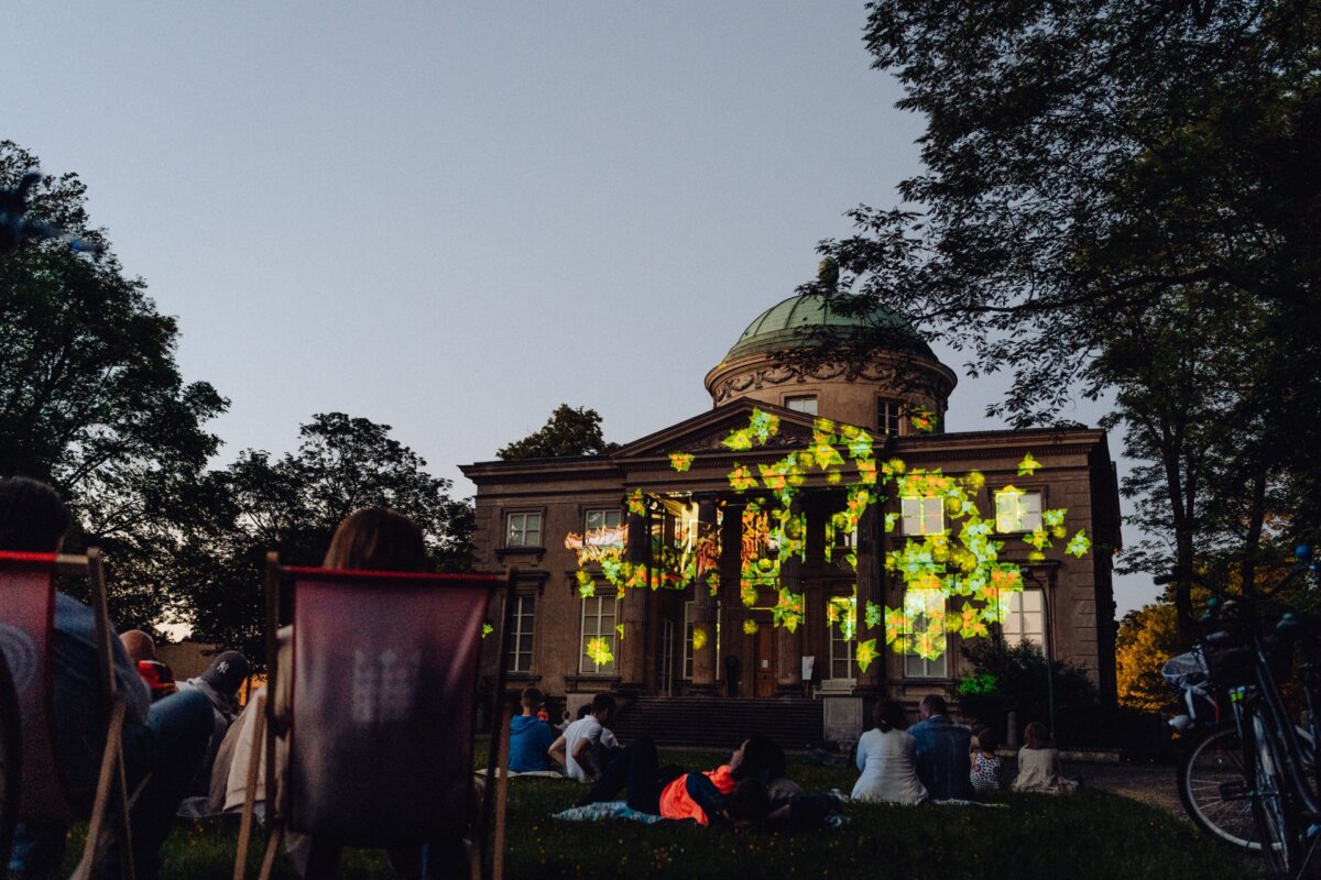 A group of people sit on the grass and in deck chairs, watching a building with a domed roof illuminated by colorful, abstract light projections. The scene is set at dusk, surrounded by trees, creating a serene atmosphere for an outdoor event - a perfect photo-op of the event. 