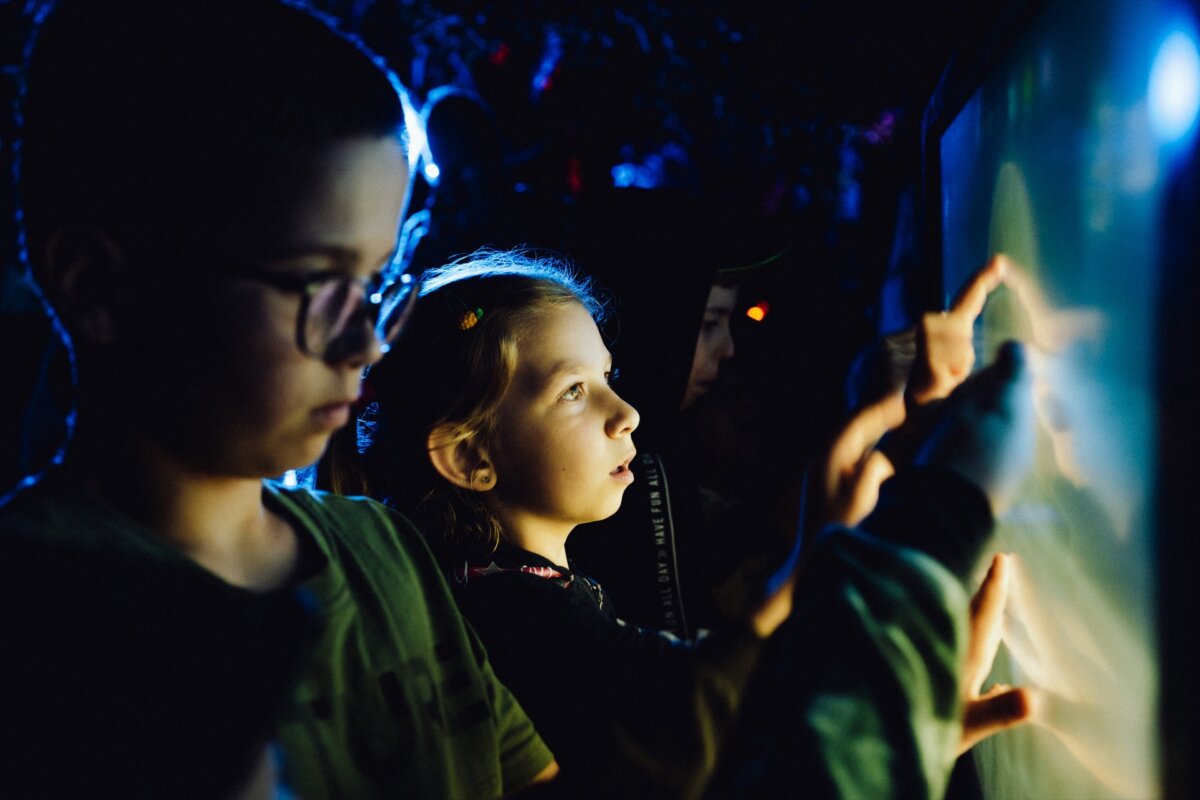 A group of children illuminated by dim blue light interact with a touch screen. The children are engrossed in what they are doing, several hands touching the screen. The background is dark, highlighting the glow of the display - perfect for a photo essay of the event.  