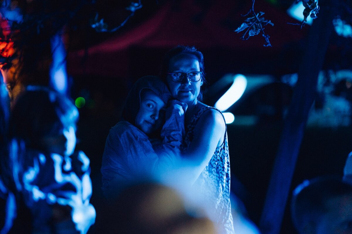 A woman and a young child embrace in blue light, creating a peaceful and comforting scene. They appear to be at an outdoor event in a dimly lit environment, with blurred silhouettes of other people in the background, capturing the essence of this event photo essay. 